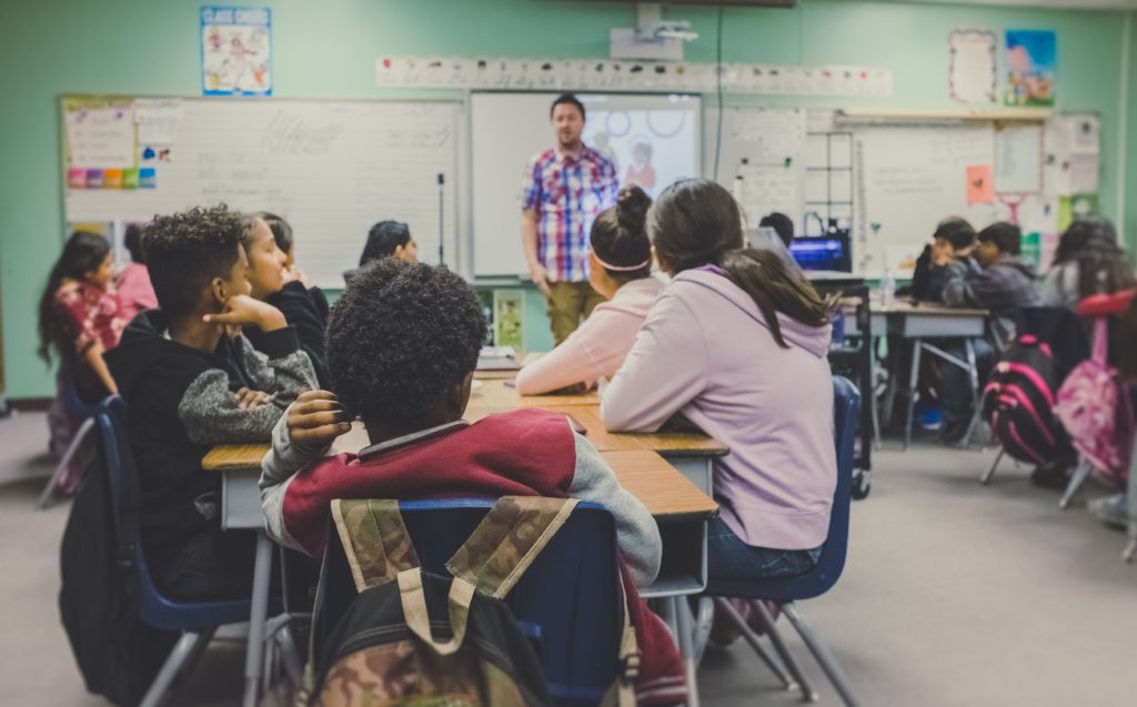 Image of a teacher in front of a secondary classroom with students engaged in discussion.